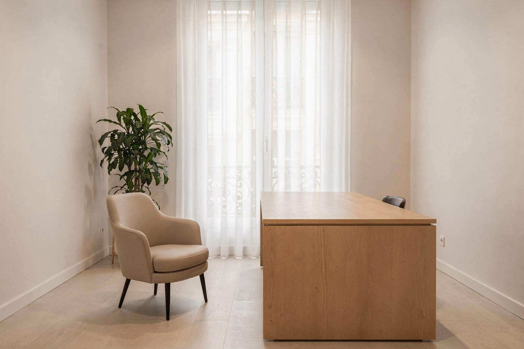 Cabinet de consultation parisien vide, bureau en chêne clair et fauteuil devant une fenêtre haussmannienne